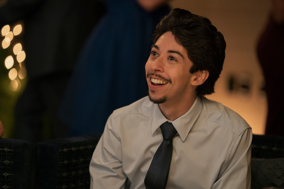 Smiling man in dress shirt and tie sitting indoors at a social event, with warm lighting and people in the blurred background.