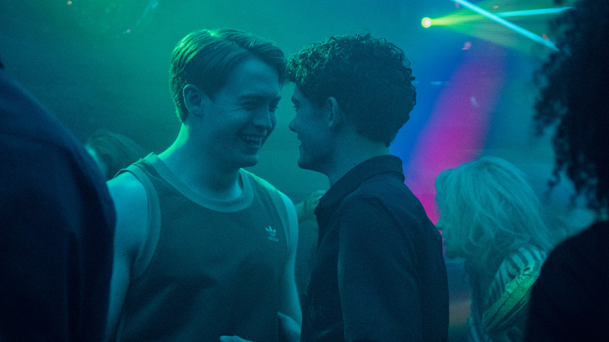 Two teenagers in a cozy, dimly lit bedroom, one sitting on the floor using a laptop, the other sitting on a bed watching. The room has string lights, posters, and personal items, creating a relaxed, lived-in atmosphere.
