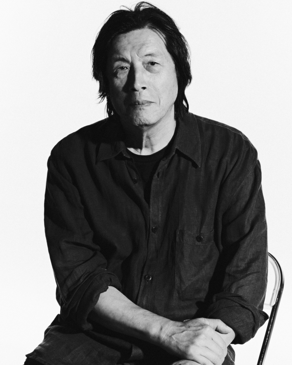 Black and white portrait of a person with medium-length hair, seated on a chair against a plain white background, wearing a dark, button-up shirt with rolled-up sleeves, looking directly at the camera.
