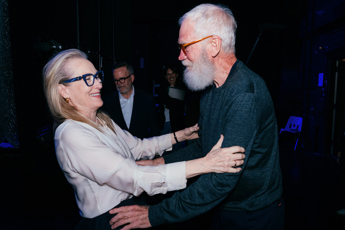 Meryl Streep and David Letterman smiling and embracing each other warmly backstage in a dimly lit setting, with a few other people and stage equipment visible in the background.