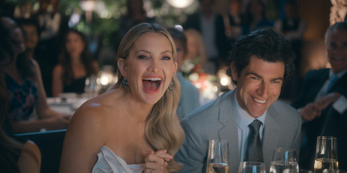 Woman in a white dress and man in a suit smiling and laughing while seated at a formal dinner event, surrounded by elegantly dressed guests and candlelit tables in a festive, celebratory atmosphere.