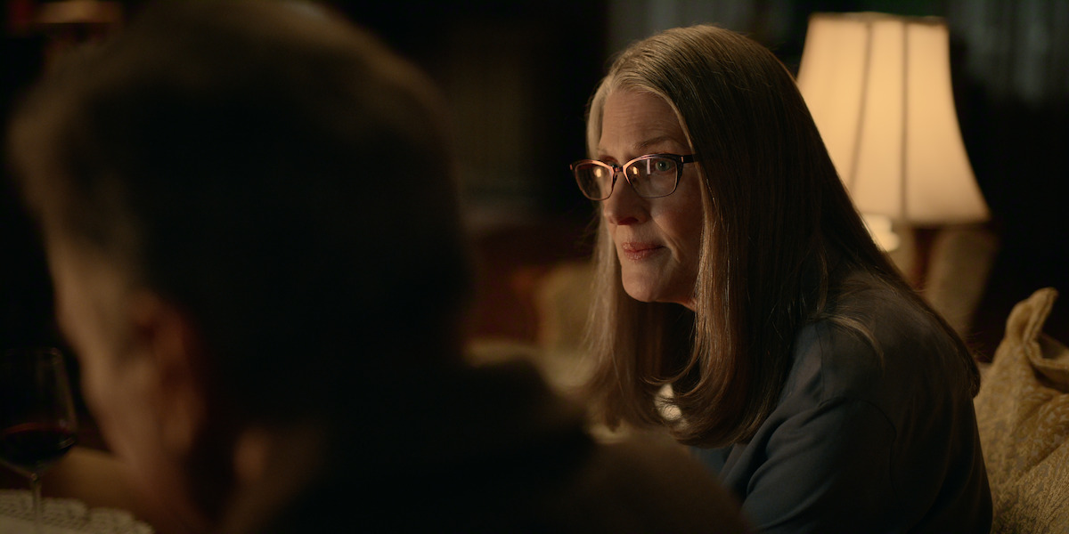 An older woman with long gray hair and glasses sits indoors on a sofa at night, softly lit by a table lamp, facing another person who is partially visible in the foreground; a cozy and intimate setting.