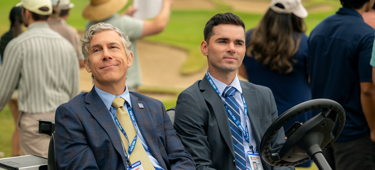 Two men in suits with lanyards sit in a golf cart at an outdoor golf course event, surrounded by people and sand traps in the background.