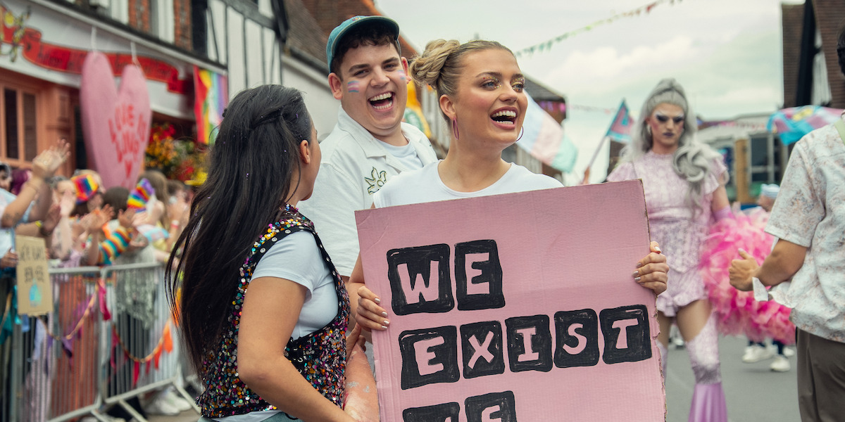 People happily marching in a pride parade on a city street, holding a pink sign that says "We exist," with colorful decorations, festive outfits, and crowds celebrating in the background.