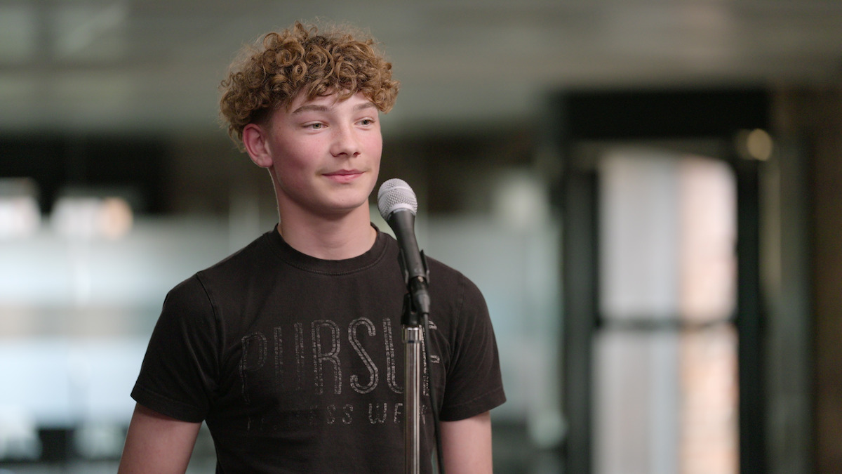 Teenager with curly hair stands at a microphone indoors, casual clothing, softly lit background, appearing confident and calm, possibly preparing to speak or perform in a relaxed, modern environment.