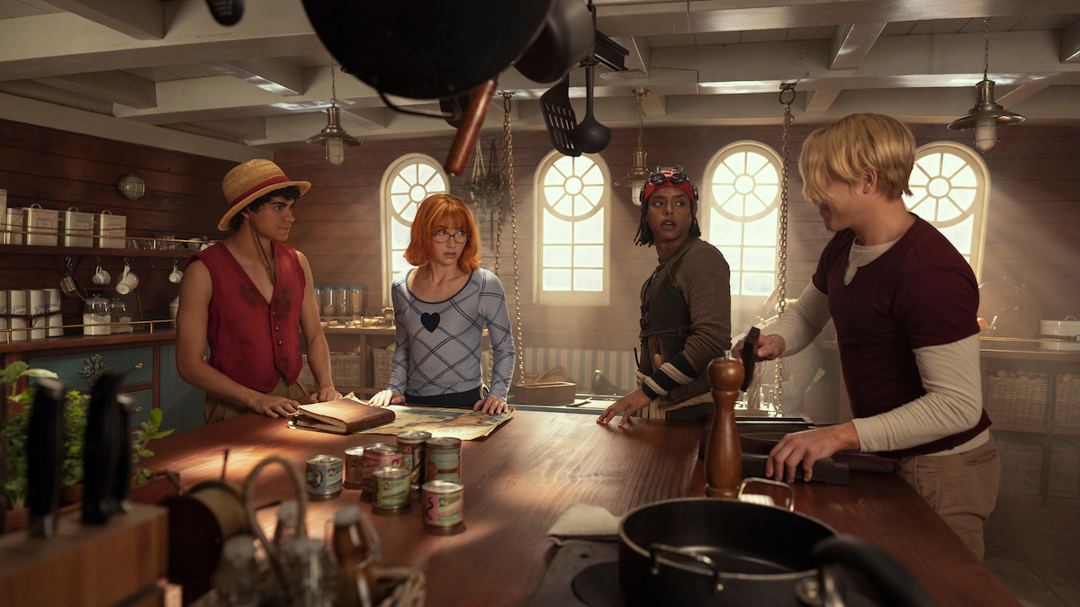 Four people in colorful clothing stand in a sunlit wooden kitchen with large windows, surrounded by pots, cans, and cooking utensils, engaged in discussion around a kitchen island.
