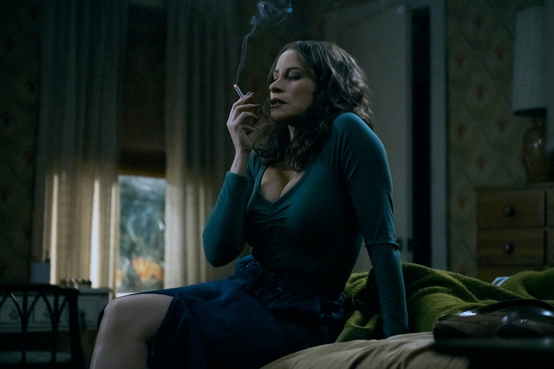 Sofia Vergara as Griselda seated on a bed, smoking a cigarette.