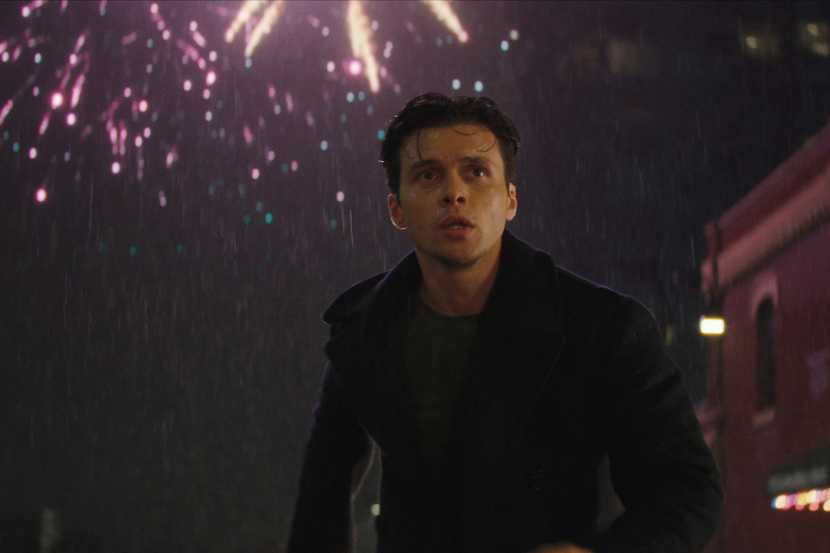 A young man in a dark coat stands outside at night in the rain, looking up as colorful fireworks explode in the sky above a city street with buildings in the background.