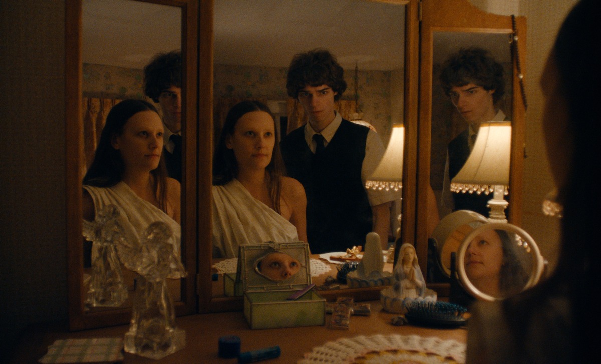 A woman in a white dress sits at a vanity with a man standing behind her. Their reflections are seen in multiple mirrors, surrounded by various decorative items and soft lamp lighting in a warmly lit bedroom.