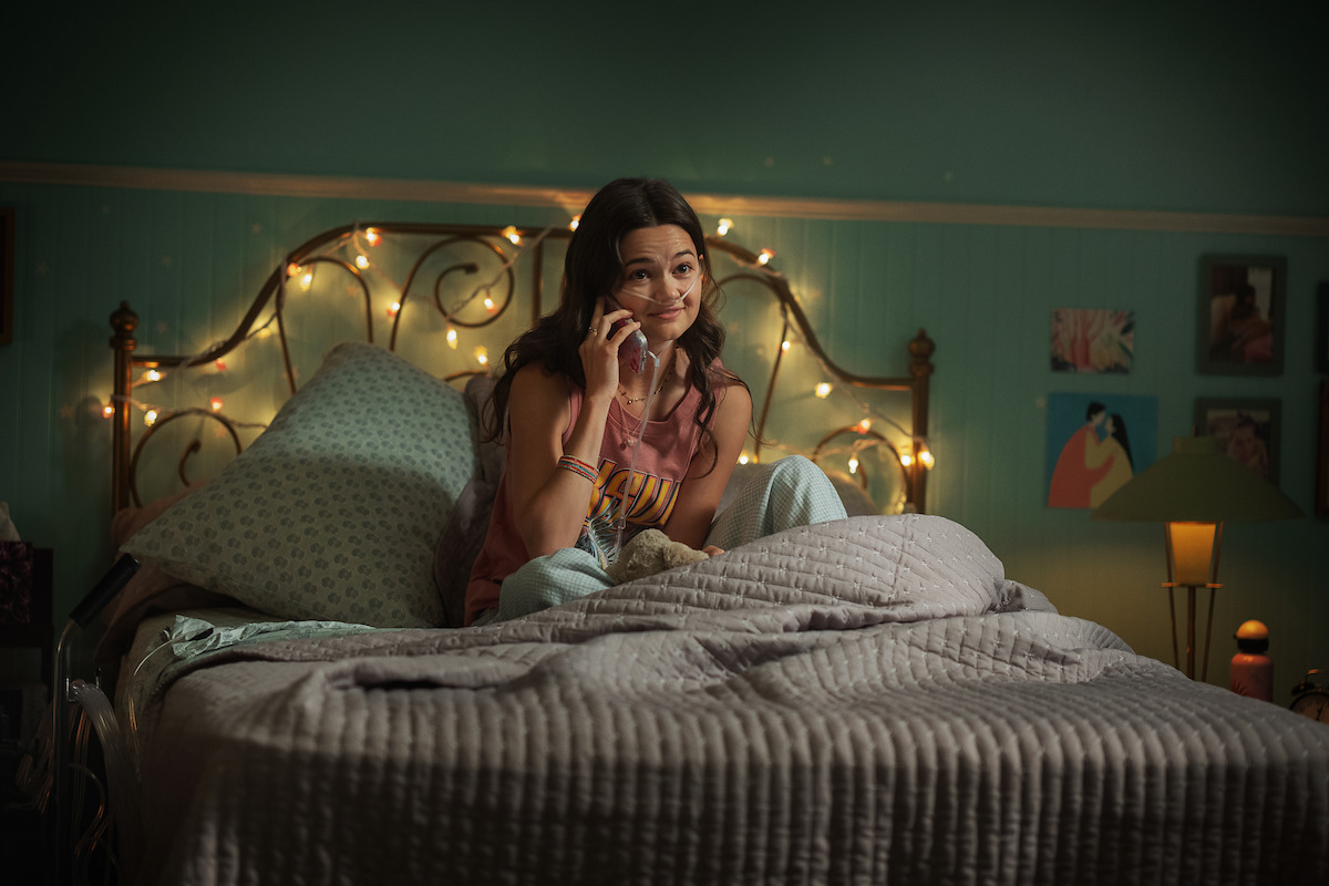 Teen girl sitting on her bed in a cozy, dimly lit bedroom, talking on the phone. The room has string lights, teal walls, pictures, and a lamp, creating a warm and relaxed atmosphere.