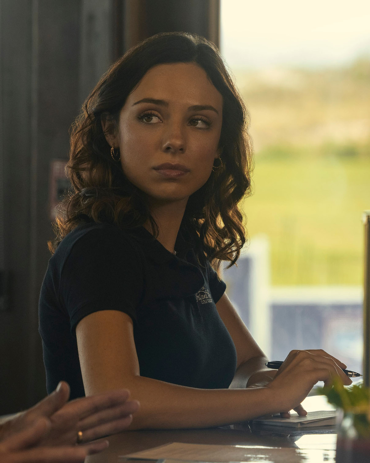 Fiona Palomo as Sofia, with wavy brown hair sits at a counter in a sunlit indoor setting, looking to the side with a thoughtful expression. The background is softly blurred with hints of greenery outside the window.