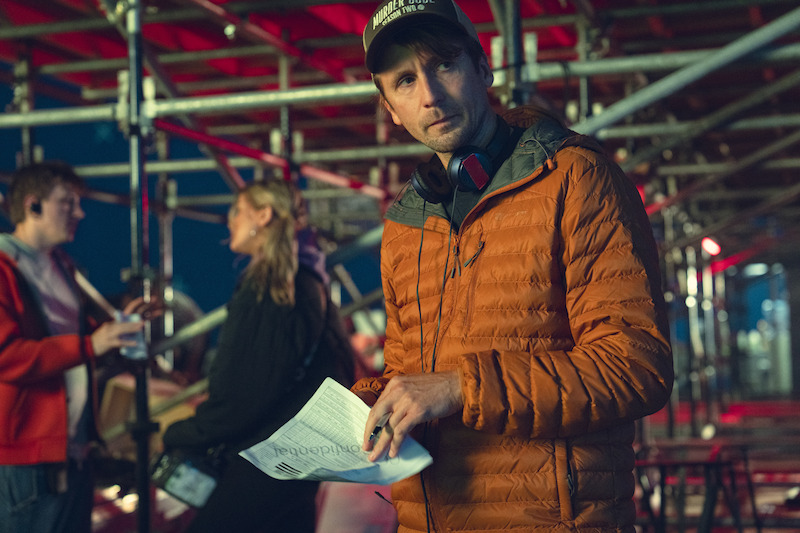 Man in orange jacket holding papers and wearing headphones stands under metal scaffolding, with people talking and working in the blurred background, suggesting a film set or production environment.