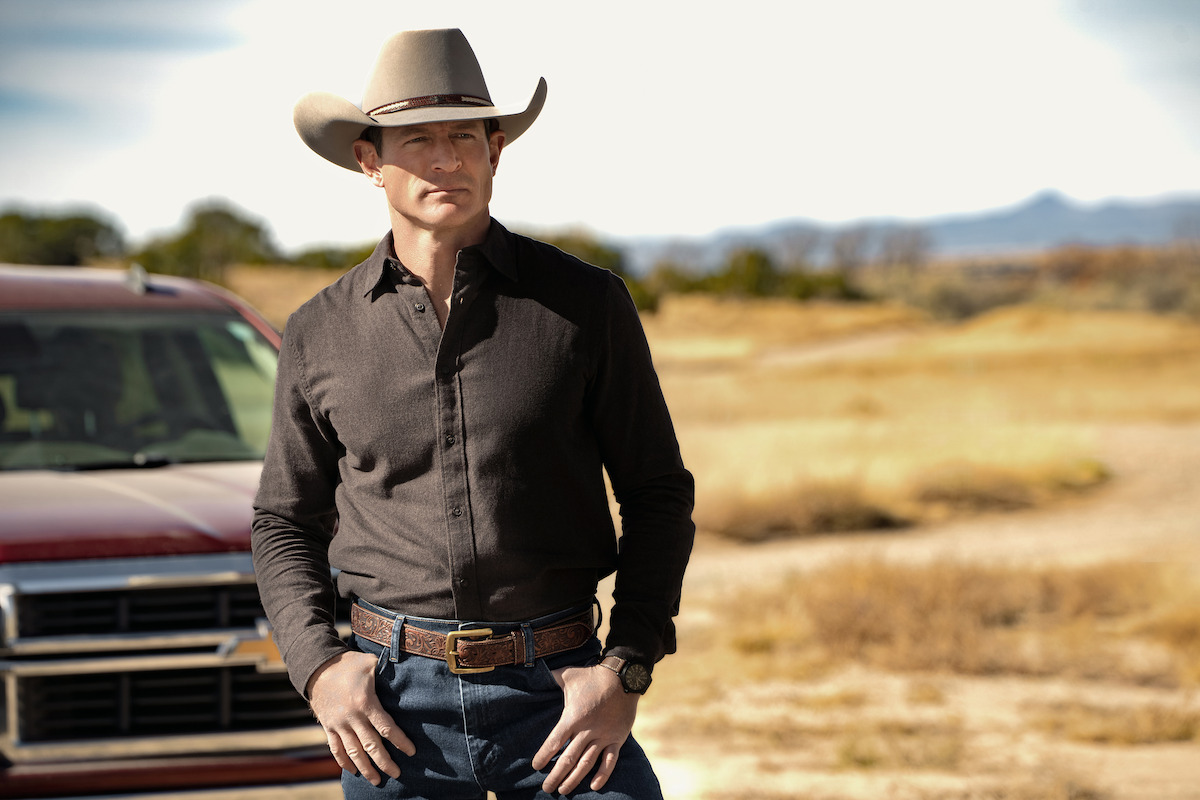 Man in cowboy hat and dark shirt standing outdoors in a rural, open field near a red truck, with distant mountains and blue sky in the background.