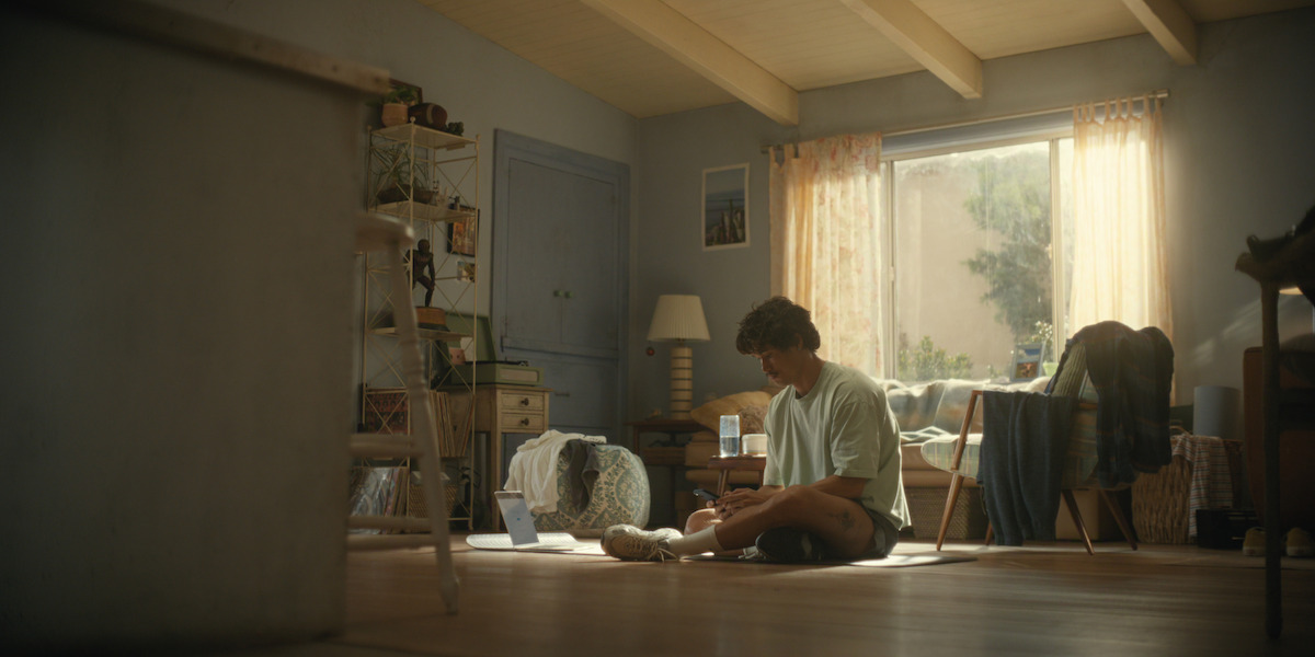 A person sits on the floor tying their shoe in a sunlit, cozy bedroom with a bed, desk, and shelves. The room is softly lit by daylight streaming through a window with light curtains, creating a calm, lived-in atmosphere.