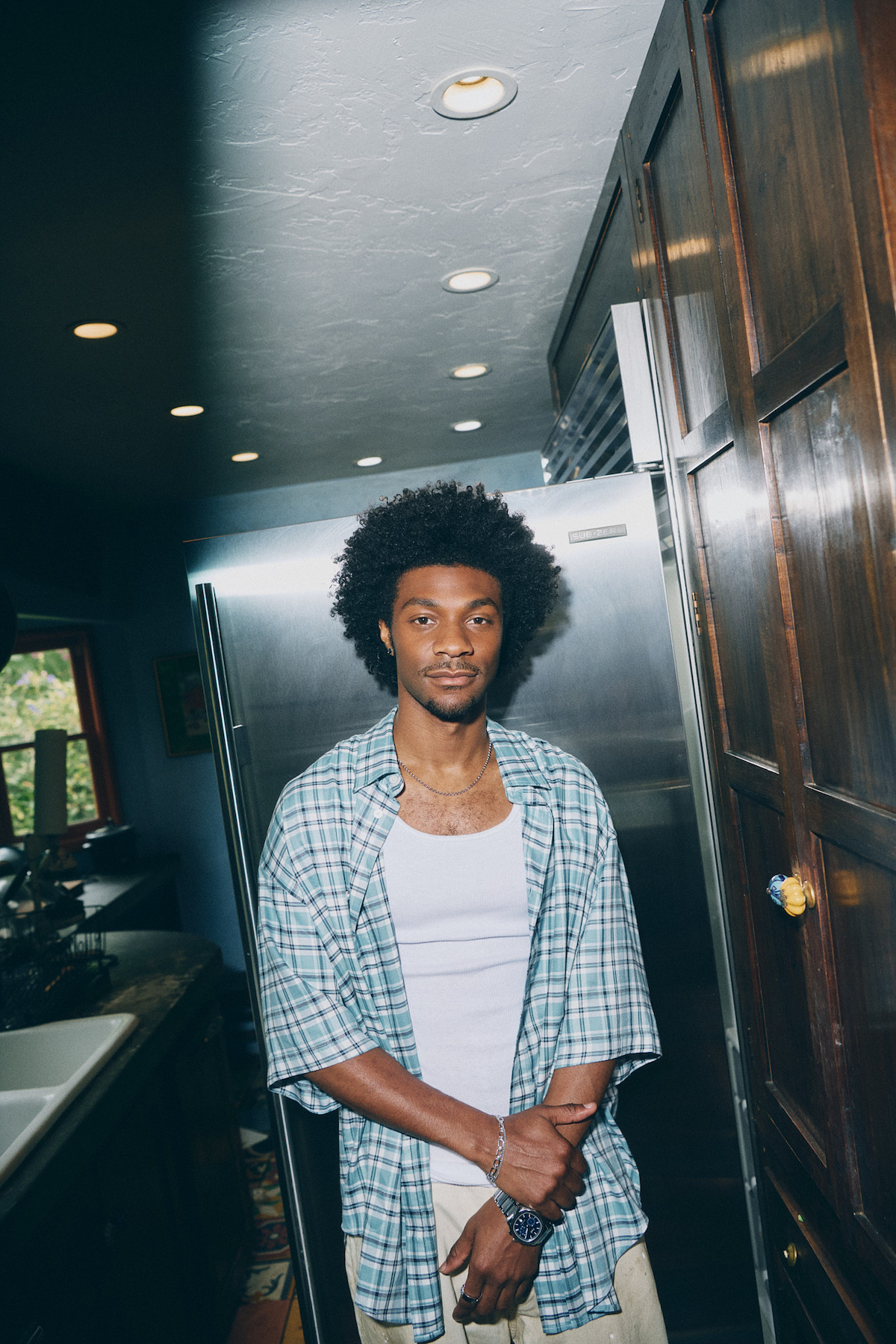 A young man with curly hair stands in a modern kitchen near a stainless steel refrigerator, wearing a plaid shirt over a white tank top. The kitchen has wooden cabinets, overhead lights, and a window letting in daylight.