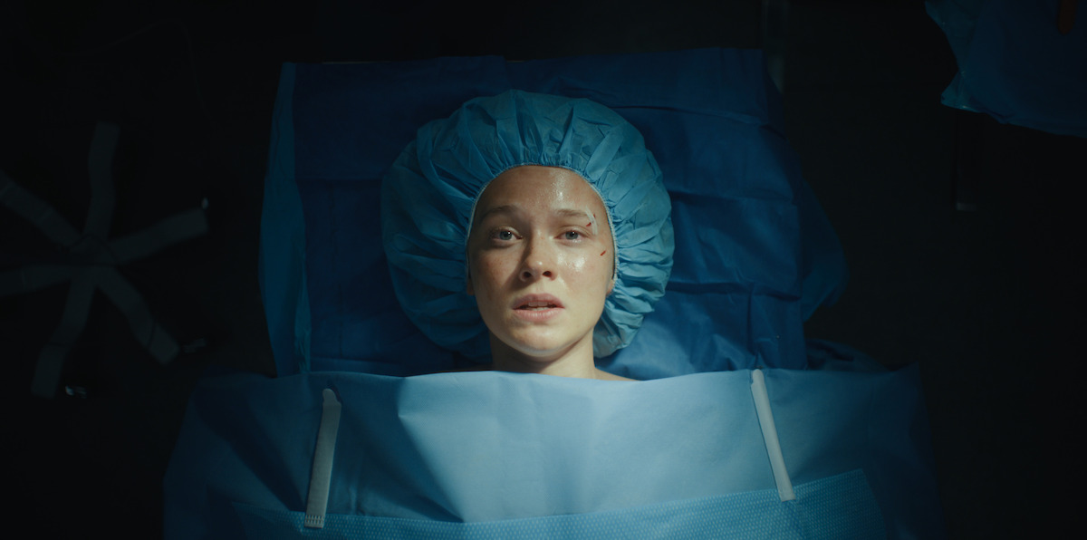 Woman lying on an operating table, covered in blue surgical drapes, wearing a surgical cap, looking up with a concerned expression, in a dimly lit hospital or surgery room.