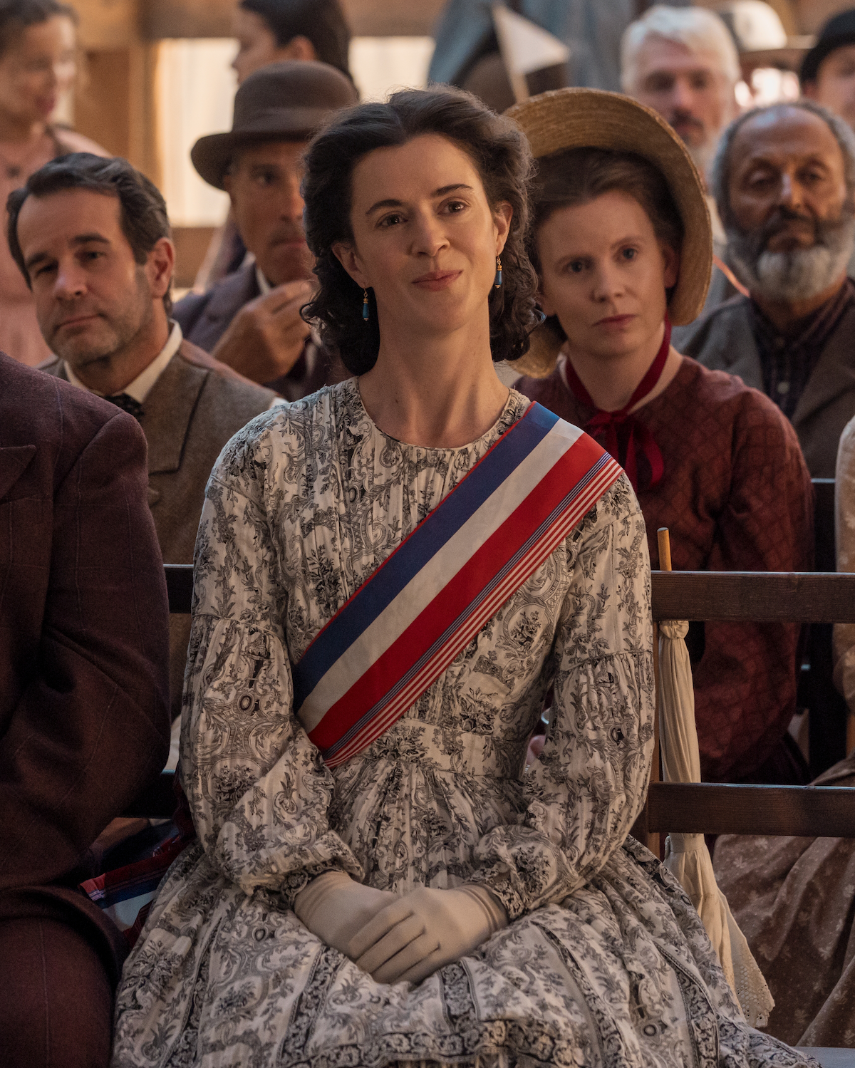 Woman in a historic patterned dress with a tricolor sash sits among others at a period event, indoors with wooden benches, dressed in 19th-century attire, focused on something ahead, suggesting a formal gathering or ceremony.