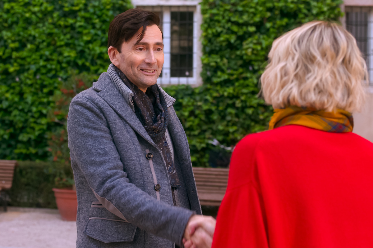Man and woman shaking hands outdoors in a courtyard with greenery and benches, both wearing coats, smiling and engaged in conversation.