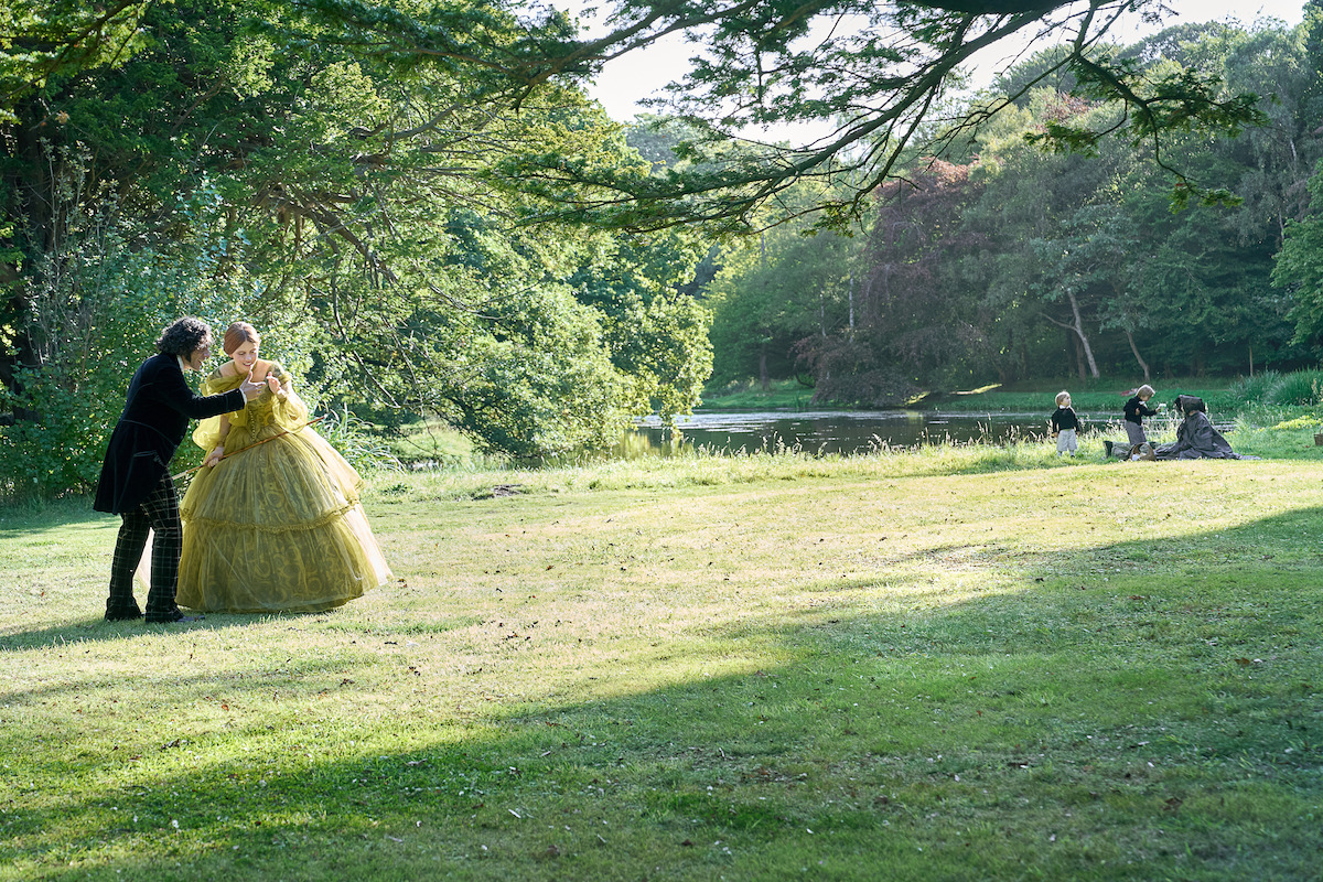 Two men in Victorian-era clothing stand in a dimly lit, wood-paneled room with framed portraits, covered furniture, and soft sunlight streaming through a window, creating a mysterious and tense atmosphere.