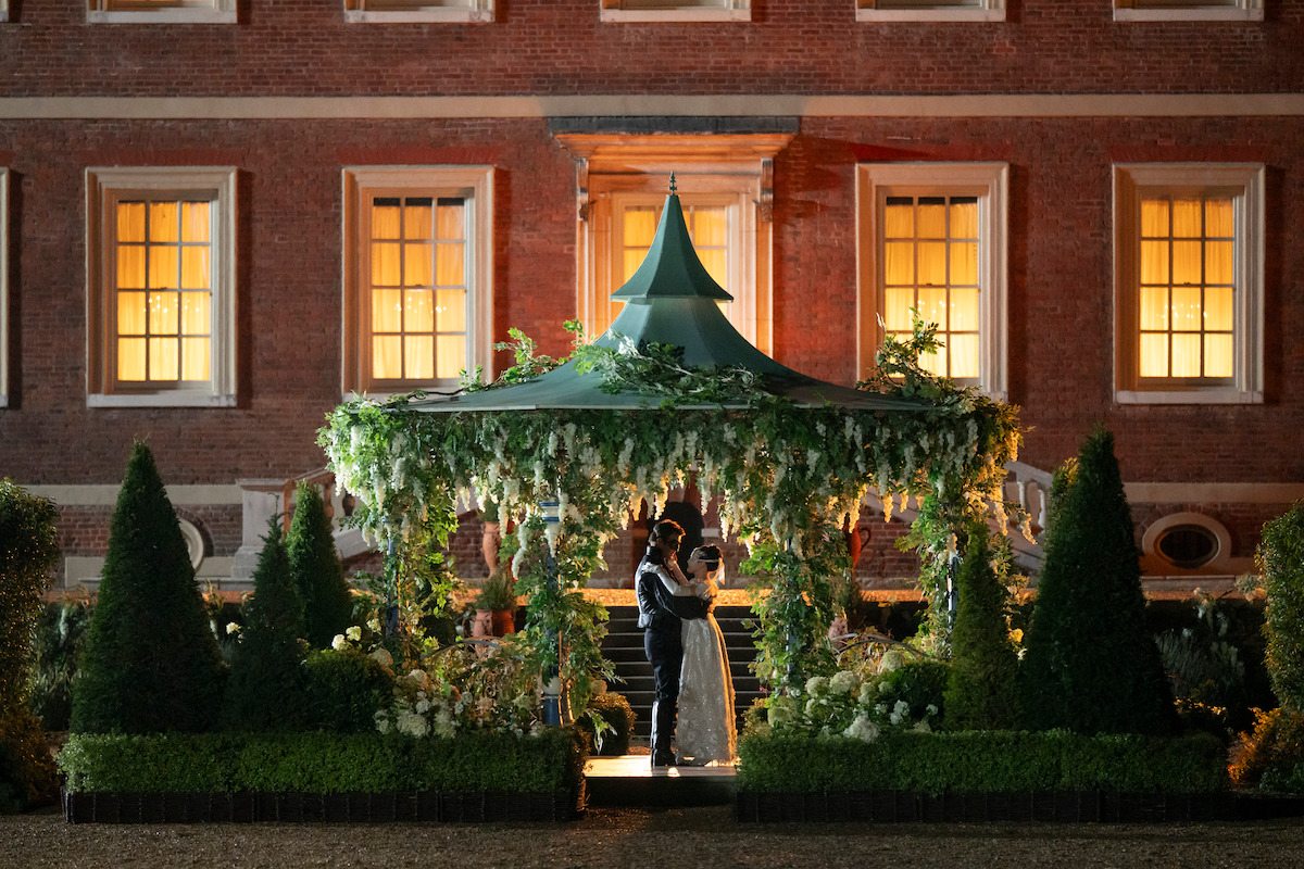 A couple embraces under a decorated, leafy canopy at night in a formal garden, in front of a large historic mansion with warmly lit windows and manicured hedges.