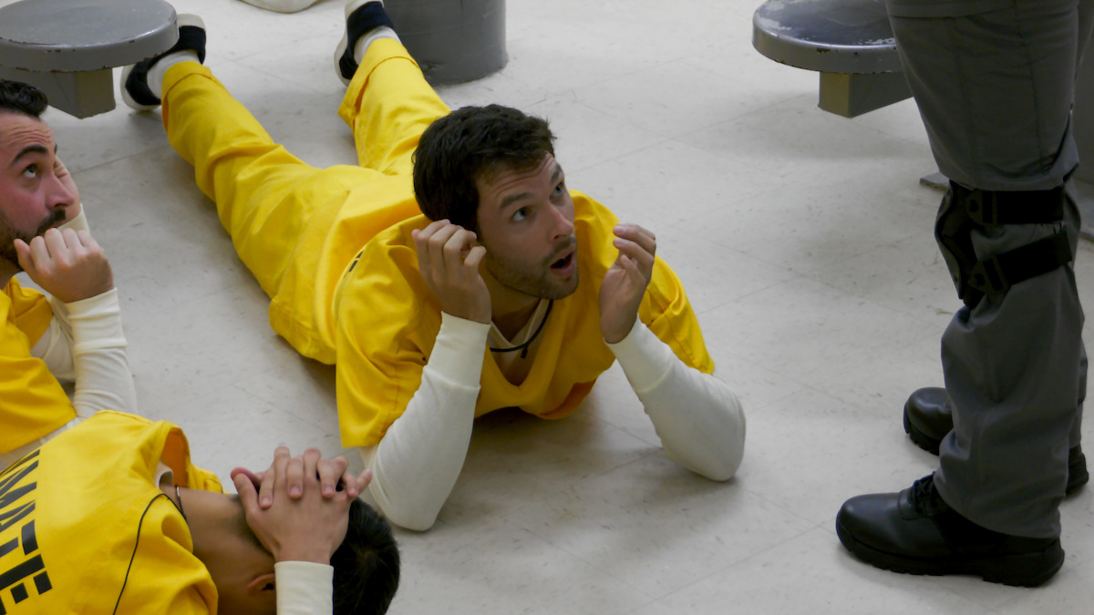 Three inmates in yellow uniforms lie on a prison floor, looking up at a standing guard in gray uniform. The environment is a correctional facility with benches and a sterile, institutional setting.