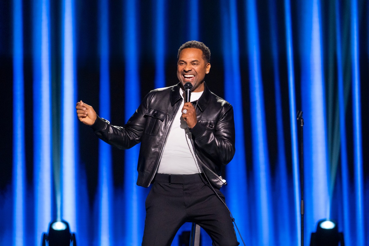 A comedian in a black leather jacket performs on stage with a microphone, smiling and animated, against a blue curtain backdrop with stage lights.