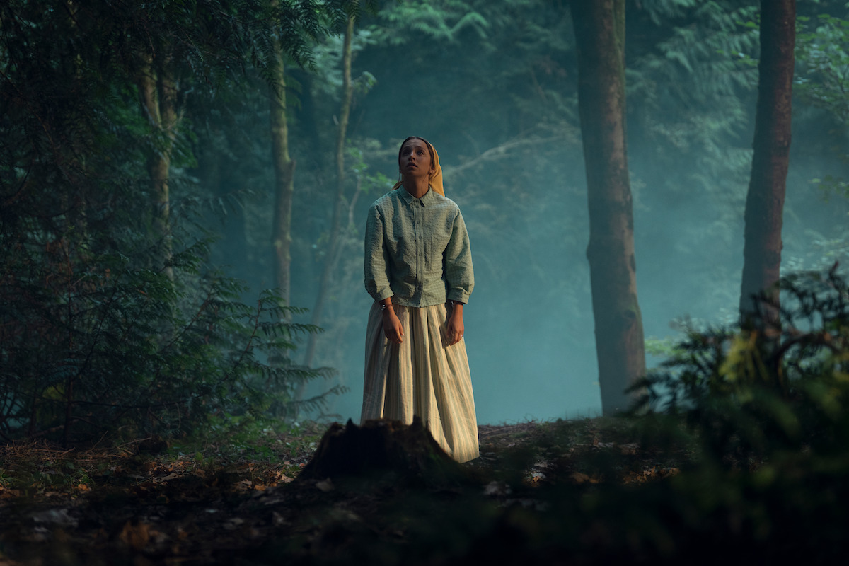 A woman in a long skirt and jacket stands alone in a misty forest, surrounded by tall trees and ferns, looking upward as soft light filters through the dense woodland environment.