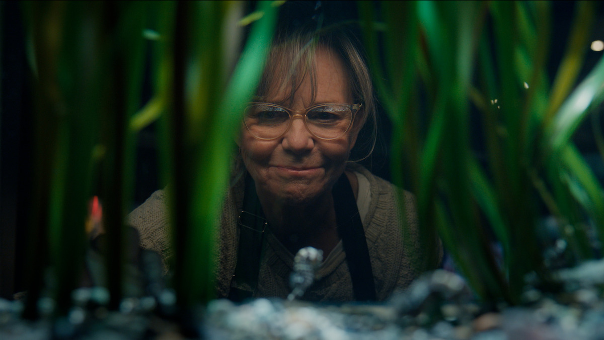 Sally Field as Tova wearing glasses looking into an aquarium tank, viewed through tall green aquatic plants and aquarium decorations, with a focused expression in a dim indoor setting.