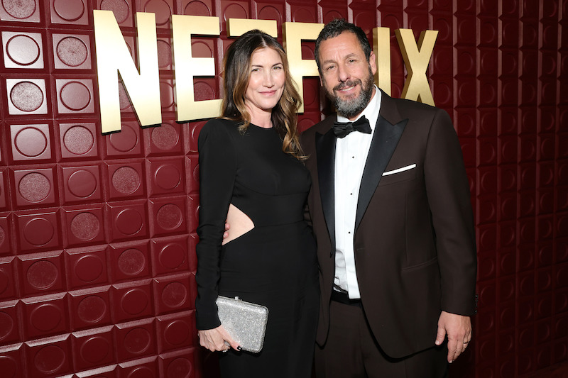 Jackie and Adam Sandler stand in front of a red backdrop.
