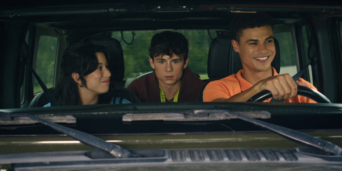 Three teenagers sitting in a car, one driving and smiling, one looking serious in the passenger seat, and one smiling in the back seat, seen through the windshield during daytime.