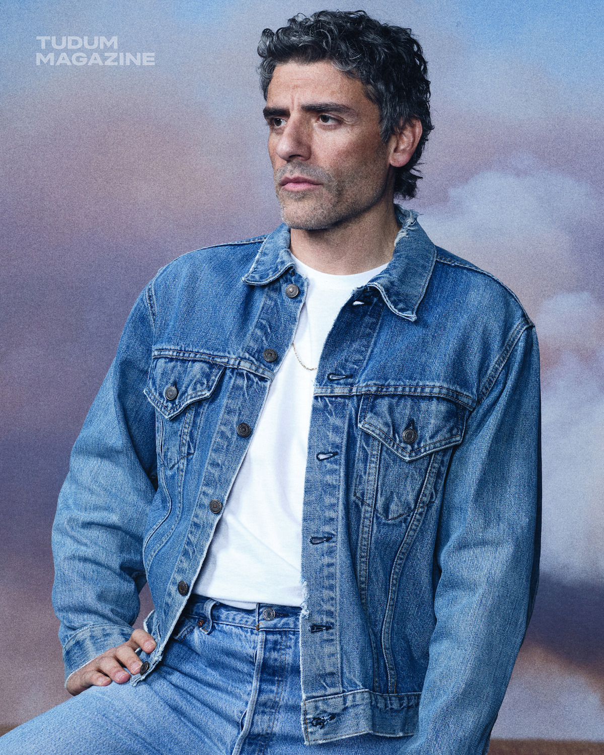 Man wearing blue denim jacket and jeans with a white t-shirt, sitting and posing in front of a cloudy sky backdrop. Studio portrait for Tudum Magazine, looks thoughtful and relaxed in a natural setting.