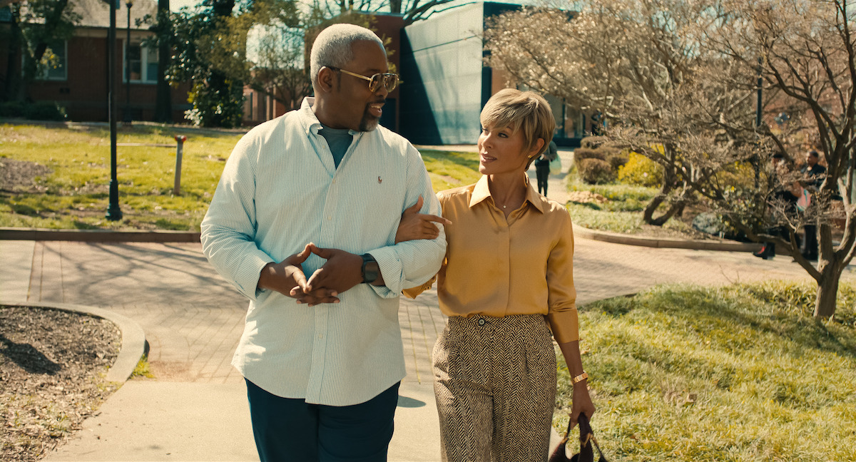 Two adults walk arm in arm along a sunny park path, surrounded by trees, grass, and sidewalks, engaging in conversation in a relaxed outdoor setting.