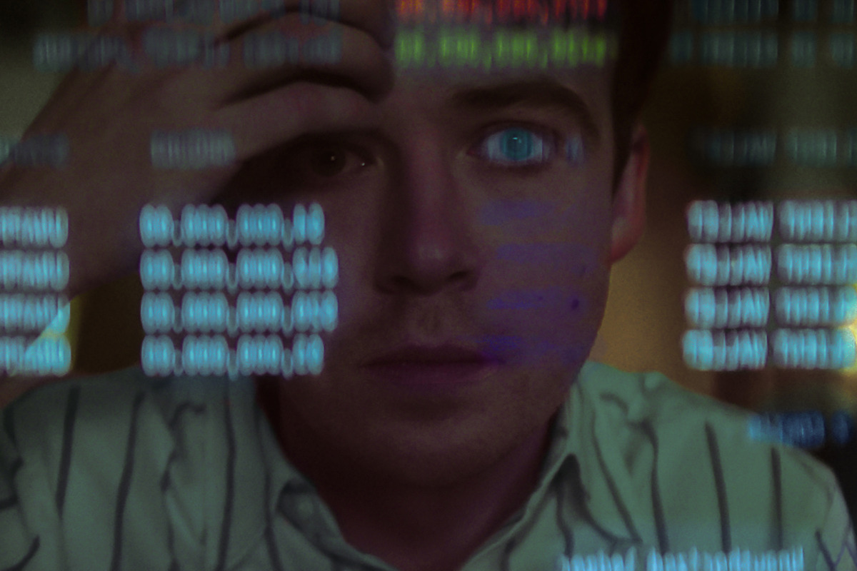 A person in a striped shirt looks intently at a computer screen, reflected data and code overlaying their face, suggesting concentration and work in a technology-focused or programming environment.
