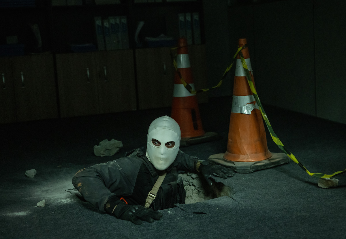 Um homem vestindo touca ninja sai de um buraco no chão de uma sala escura