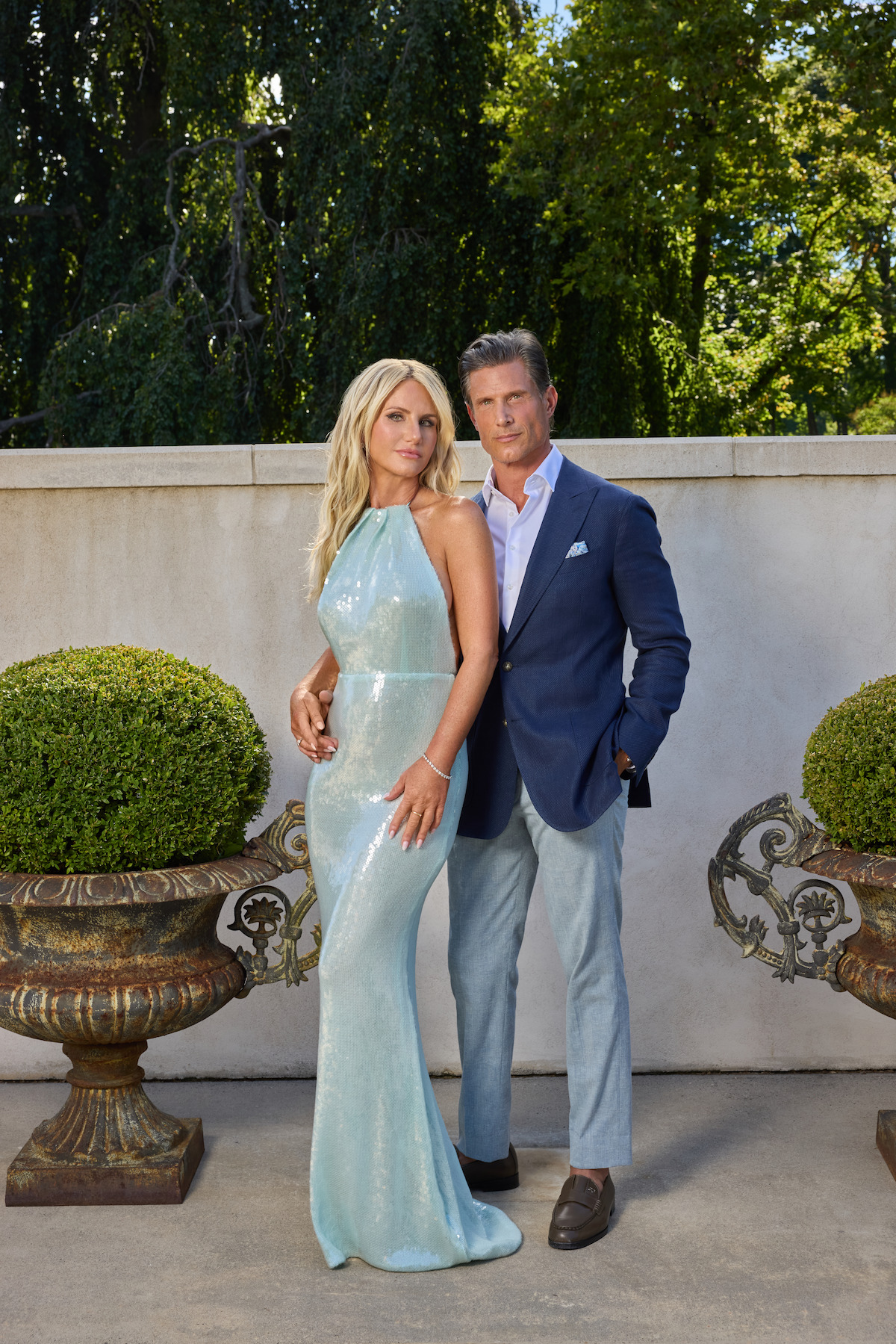 A well-dressed couple poses outdoors between two large ornate planters, with trimmed bushes and a stone wall behind them, surrounded by lush green trees in the background.