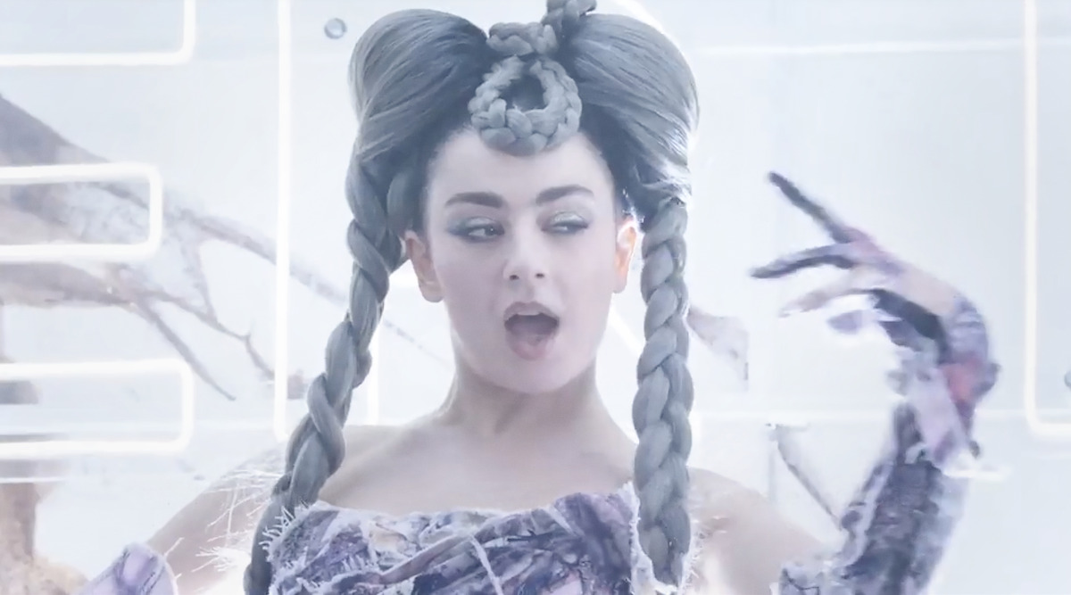 Person with elaborate braided hair and unique outfit poses in a futuristic, brightly lit environment with abstract elements in the background.
