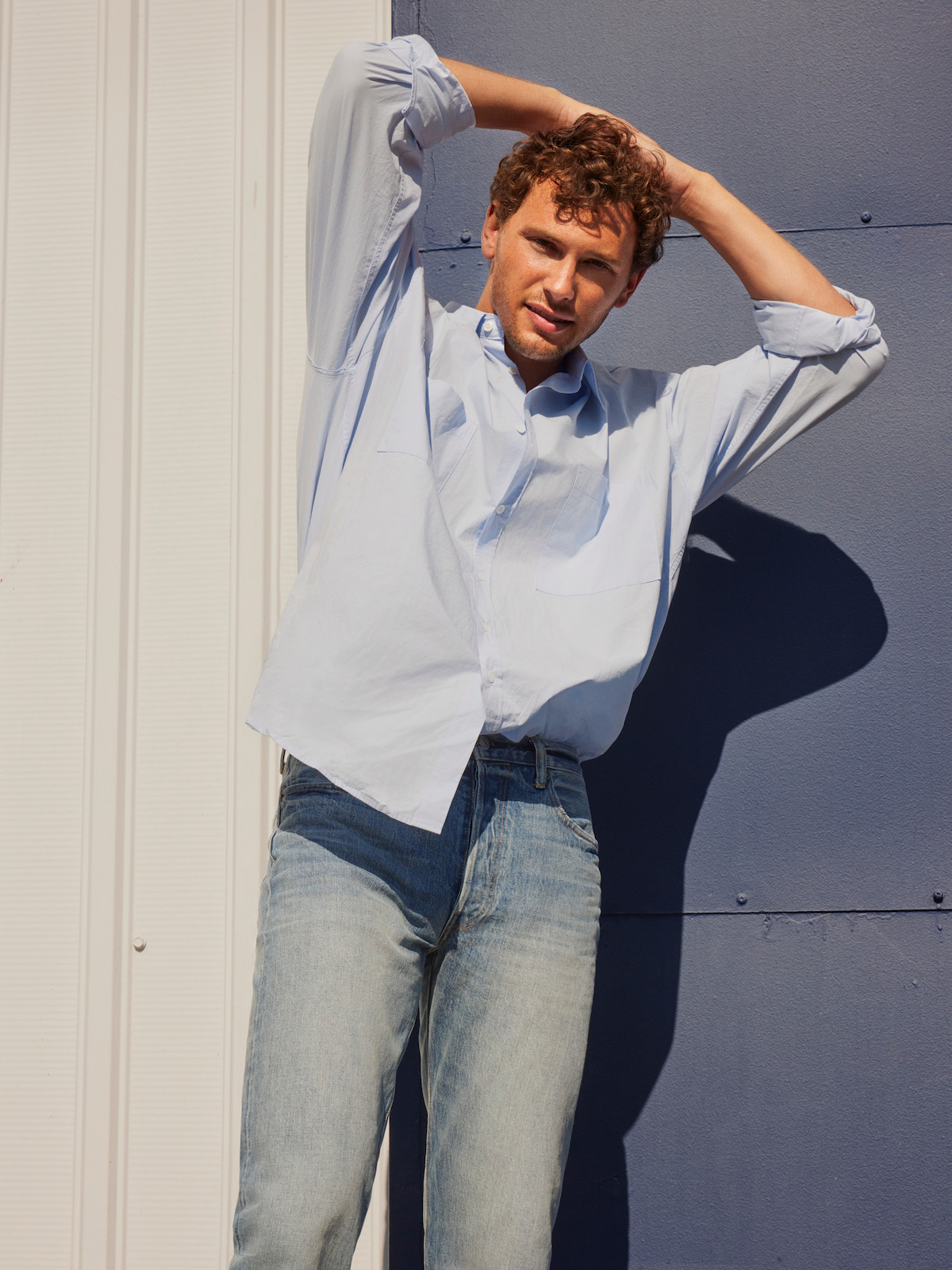 Cooper Koch looking into the camera, posing with his hands over his head wearing a blue button down and jeans.