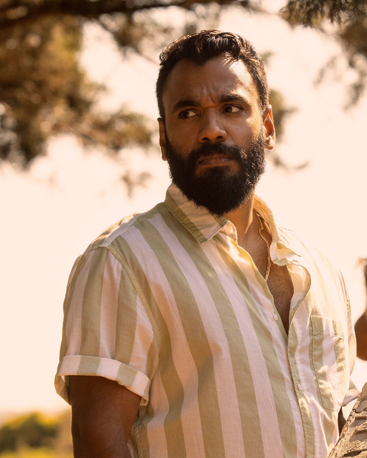 Andy McQueen as Carlos Singh, with a beard wearing a light green and white striped short-sleeve shirt stands outdoors in warm sunlight, surrounded by blurred trees and natural greenery in the background.