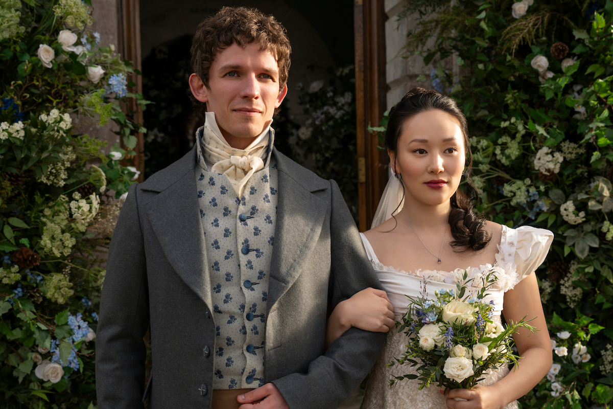 A man and woman in period attire stand arm in arm outside, surrounded by flowers and greenery, at a wedding ceremony. The bride holds a bouquet and both appear serene and elegant.
