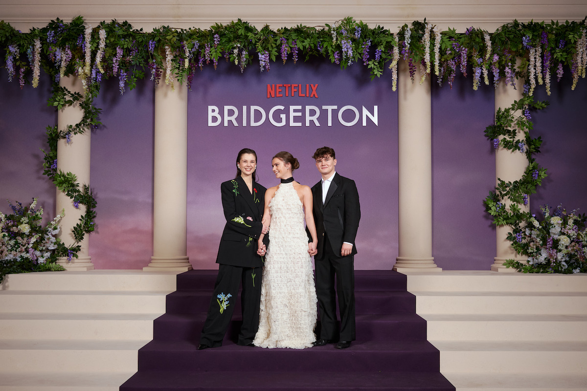 Three people pose on purple carpeted steps in formal attire at a Bridgerton Netflix event, with floral decorations, columns, and a backdrop featuring the show's logo.