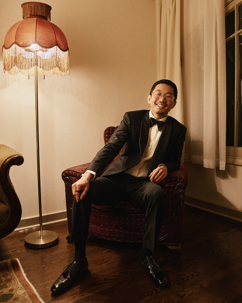 Man in a black tuxedo sits smiling on a red armchair in a warmly lit room with wooden floors, next to a vintage standing lamp and a window with white curtains.