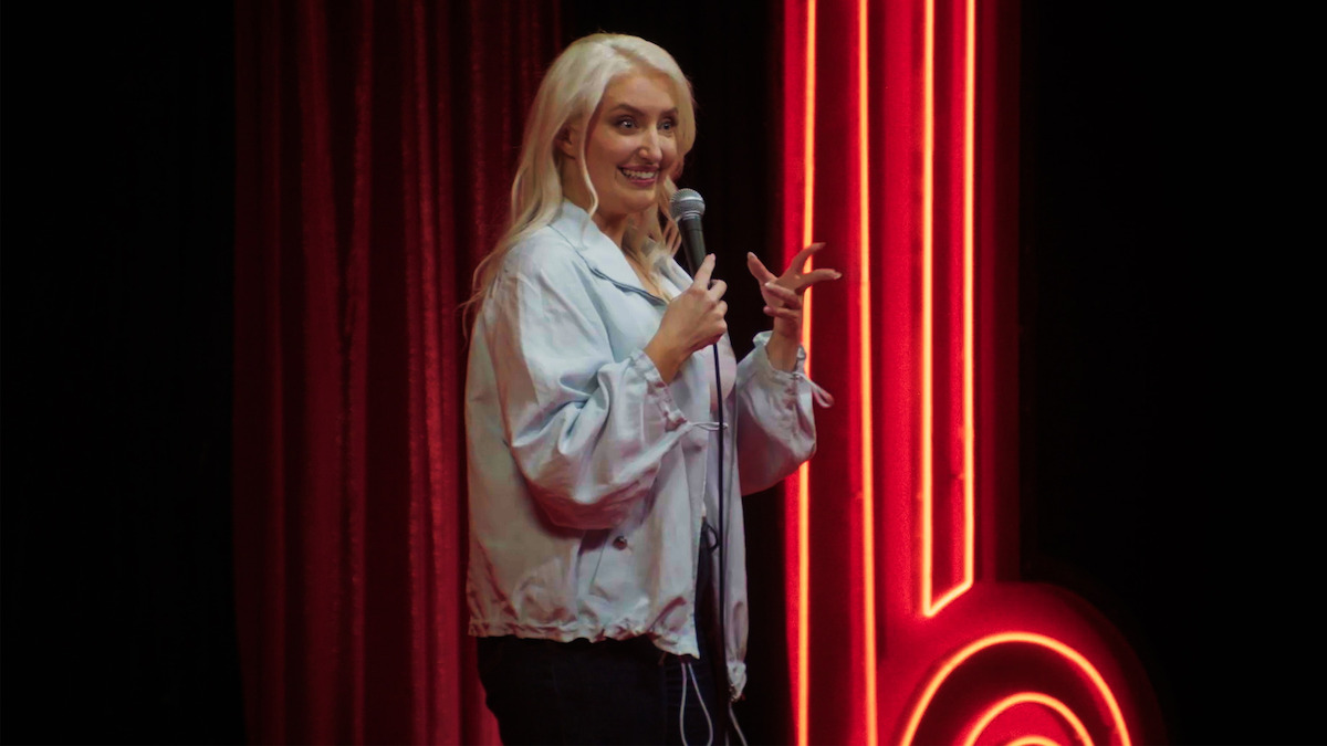 A woman with long blonde hair performs stand-up comedy on stage, holding a microphone, with a red neon light and curtain backdrop.