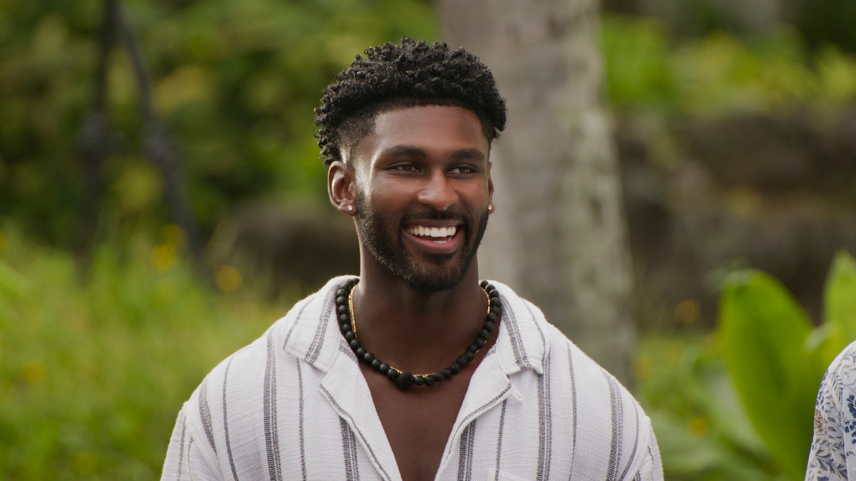 Smiling man with short curly hair and a bead necklace in a striped shirt standing outdoors in a lush, green garden with tropical plants and a tree in the background.