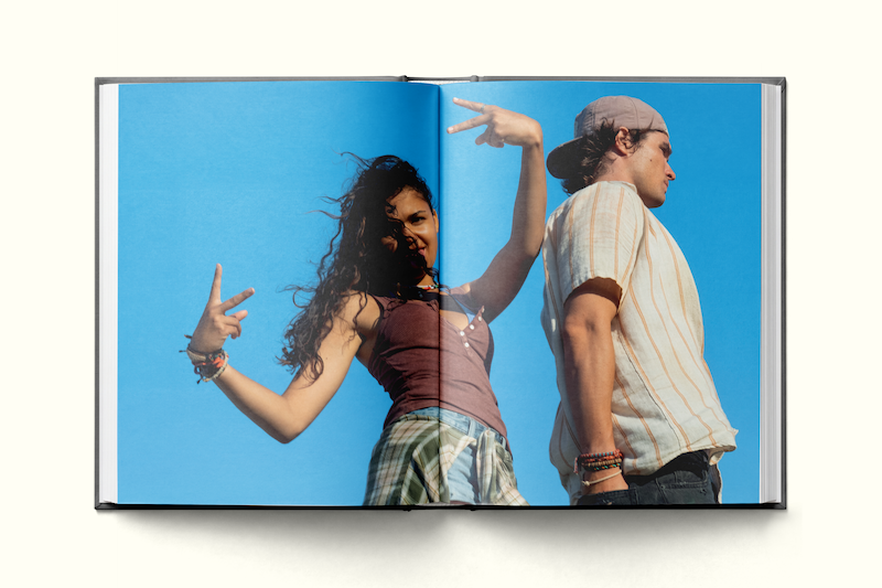 Two young adults pose playfully outdoors against a clear blue sky, standing back to back. The woman smiles and makes peace signs with her hands, while the man looks off to the side. Image is printed in an open book.
