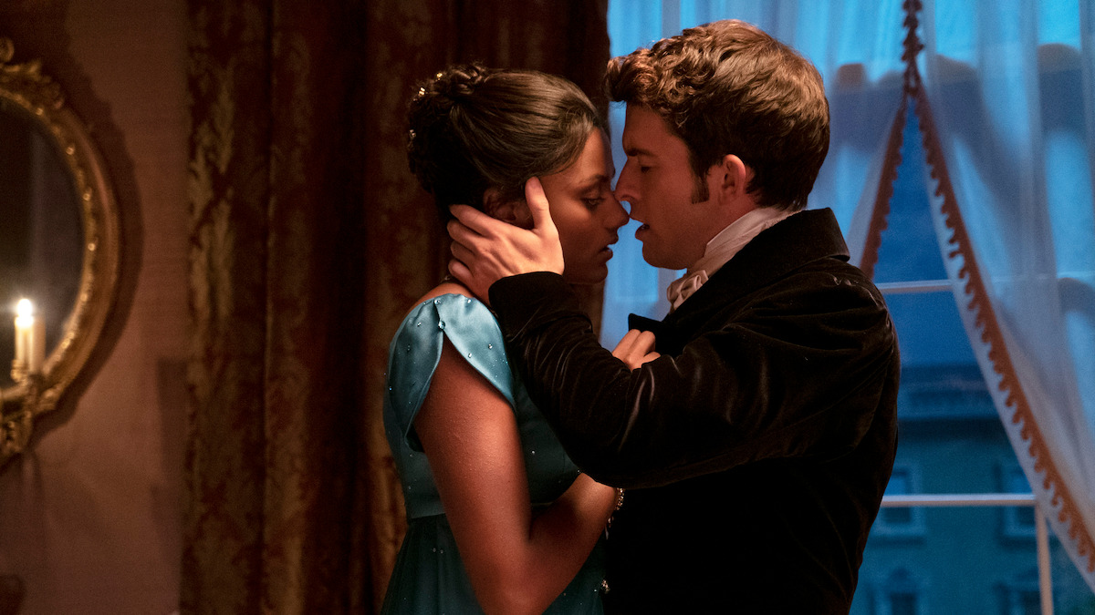 A couple in period clothing stands close together in a romantic, candlelit room with draped curtains and a mirror, about to kiss, conveying intimacy in a historical setting.