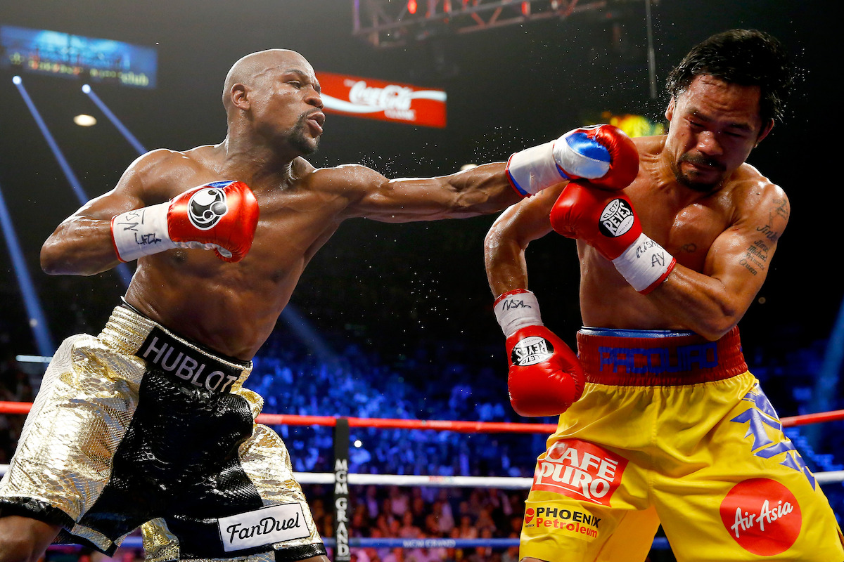 Floyd Mayweather Jr. and Manny Pacquiao are fighting in a brightly lit boxing ring surrounded by a crowd, with one landing a punch on the other's face, both wearing gloves, shorts with logos, and surrounded by blue and red ring ropes.