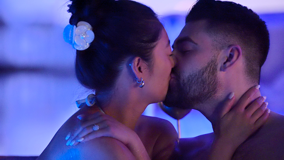 A couple sharing a passionate kiss in a softly lit, romantic setting with blue and purple lighting, a wine glass in the background, and a flower in the woman’s hair.