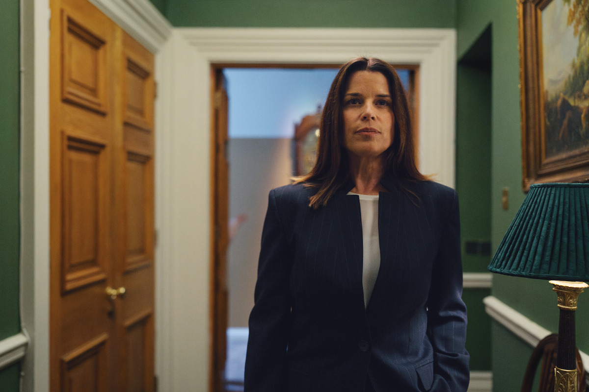 Mulher de terninho escuro parada num corredor de paredes verdes, com portas de madeira, uma luminária verde e alguns quadros. Ela tem uma expressão séria e está em um espaço formal e bem iluminado.