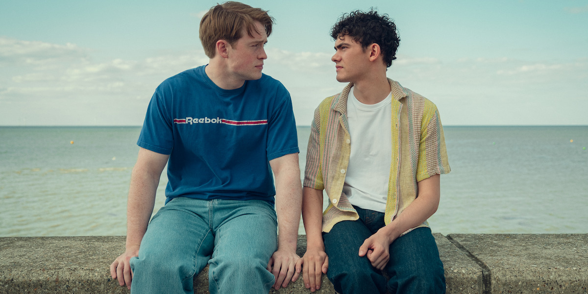 Two young men sitting on a stone wall by the sea, facing each other and holding hands, with a calm ocean and cloudy sky in the background, suggesting a moment of connection and intimacy in a coastal outdoor setting.