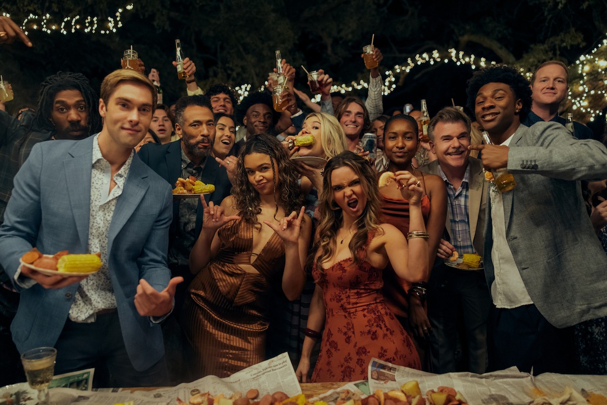 Group of young adults at an outdoor night party, dressed in semi-formal attire, holding drinks and food, smiling and posing enthusiastically with string lights decorating the background.